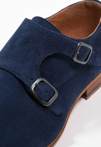 Chaussure en daim bleu foncé avec deux boucles métalliques argentées, bout arrondi et semelle en bois marron contrastante. Texture lisse avec des coutures soignées.