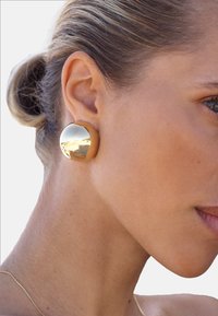Guldtonad rund studörhänge med en slät, reflekterande yta. Designen är minimalistisk, med en polerad finish och subtila kurvor.