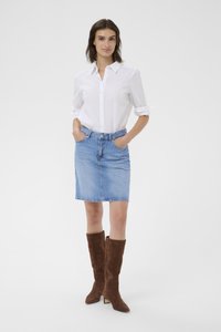 Bela srajca z gumbi, svetlo modra jeans krila, rjavi suede škornji do kolen s koničastim nosom in zmernim peto; model ima roke v žepih.