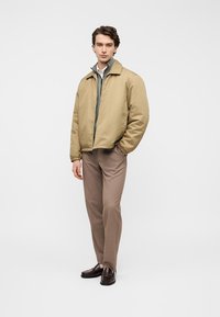 Δερμάτινο καφέ-φωτεινό bomber μπουφάν με λεία υφή, εσωτερικό γκρι στρώμα με φερμουάρ, σε συνδυασμό με κομψό μπεζ παντελόνι και σκούρα καφέ παπούτσια.