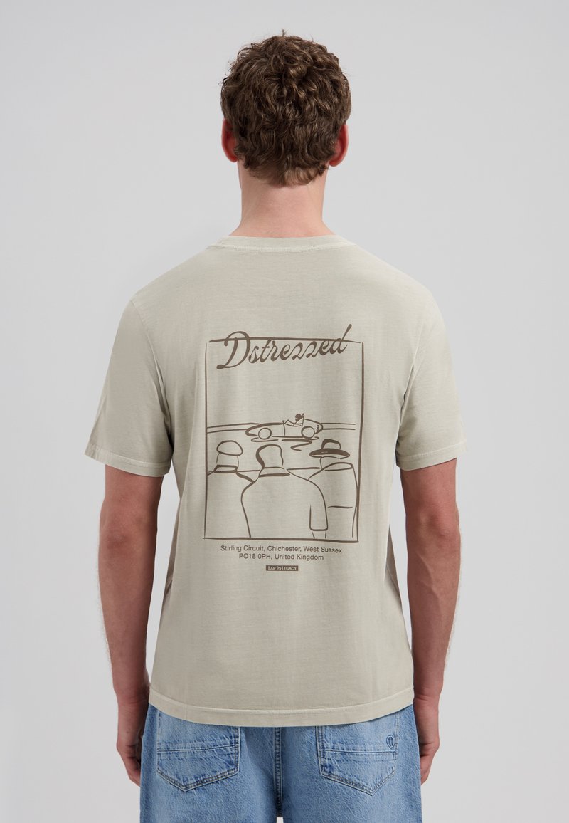 Homme portant un t-shirt beige avec un dessin linéaire de trois personnes regardant une voiture de course, texte "Dstressed" et emplacement du circuit de Stirling au dos.