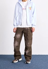 Lichtblauwe hoodie met rits, wit T-shirt met grafisch ontwerp, bruine cargobroek met meerdere zakken en bruine sportschoenen.