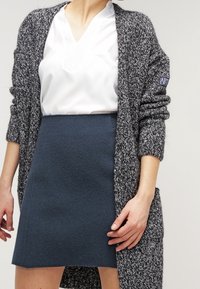 Cardigan lungo grigio e nero lavorato a maglia con apertura frontale, tessuto testurizzato e un logo discreto sulla manica, abbinato a una camicetta bianca e una gonna scura.
