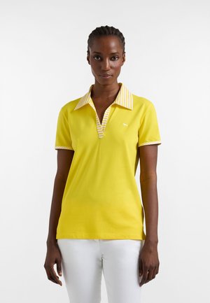 Mujer con cabello trenzado que lleva una camisa polo amarilla de manga corta con cuello y bordes de las mangas a rayas, posando frente a un fondo blanco.