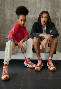 Sneaker alte con design rosso e bianco, tessuto testurizzato, suola in gomma e dettagli a zigzag. Modello che indossa pantaloni bianchi strappati e un maglione vivace.