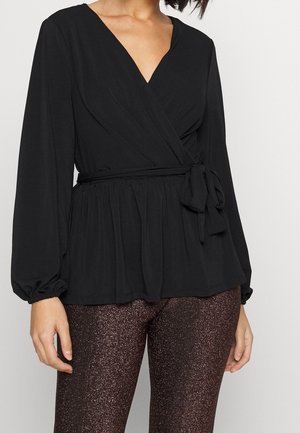 Mujer con blusa cruzada de manga larga negra con lazada en la cintura y pantalones ajustados marrón oscuro con brillo.