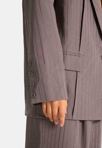 Chaqueta de traje gris con rayas verticales, adornada con pequeños strass, que presenta un puño de un solo botón y un solapa en pico.