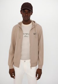 Sudadera beige con cremallera y capucha ajustable, camiseta blanca debajo, combinada con pantalones blancos. La camiseta de manga corta presenta texto impreso.