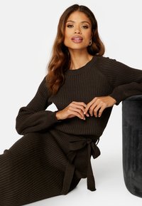 Maglione a coste marrone scuro con maniche lunghe a sbuffo, scollatura tonda e dettaglio con cintura in vita, abbinato a una gonna coordinata.