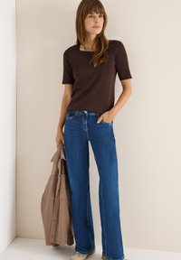 Braunfarbener Kurzarm-Top mit quadratischem Ausschnitt, kombiniert mit hellblauen Schlagjeans. Das Model hält eine beige Jacke und präsentiert ein lässiges Outfit.