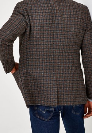 Homme portant un blazer à carreaux marron et gris avec la main gauche dans la poche, associé à un jean bleu foncé, vu de dos.