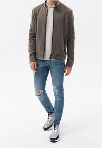 Mężczyzna w brązowej, strukturalnej kurtce na zamek błyskawiczny, białej koszulce, podartych niebieskich skinny jeansach i szaro-czarnych sneakersach na białym tle.