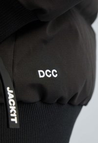 Sort træningsoutfit med tekstureret stof, der har hvid broderet skrift "DCC" og et mærketag "JACK1T." Glatte og ribbede sektioner.