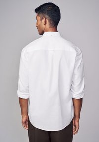 Camicia bianca a maniche lunghe con maniche arrotolate, dettaglio con piega sul retro e orlo arrotondato. Tessuto liscio e leggero. Indossata sopra pantaloni scuri.