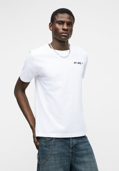 Νεαρός άνδρας με λευκό t-shirt "Off-White", αλυσίδα λαιμού και μπλε τζιν με ασημένιο αξεσουάρ αλυσίδας, στέκεται μπροστά σε απλό φόντο.