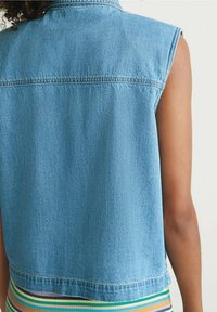 Débardeur en denim bleu clair avec une coupe droite, des détails de couture visibles et un ourlet bande sur un vêtement rayé multicolore.
