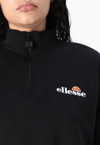 Musta vetoketjullinen collegepusero, jossa on korkea kaulus, valkoinen kirjailtu logo ja värikäs koriste. Sileä pinta ja istuva malli.