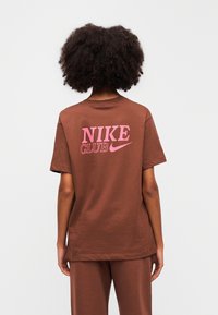 Καφέ βαμβακερό μπλουζάκι με ροζ λογότυπο «NIKE CLUB» και το σήμα Swoosh στην πλάτη. Κοντά μανίκια, άνετη εφαρμογή, ομαλή υφή.