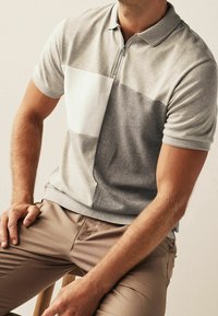 Polo gris avec une fermeture éclair à mi-longueur, panneau texturé blanc sur la gauche et manches courtes. Stylé avec un pantalon beige.