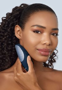 Un appareil de soin de la peau manuel avec un design ergonomique bleu foncé et une surface texturée grise, tenu contre la joue près de la ligne de la mâchoire.