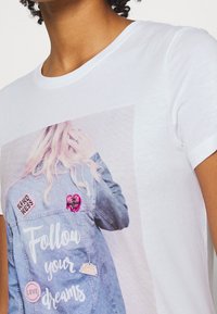 Camiseta blanca con una imagen impresa de una chaqueta de mezclilla con varios parches coloridos y el texto "Sigue tus sueños."