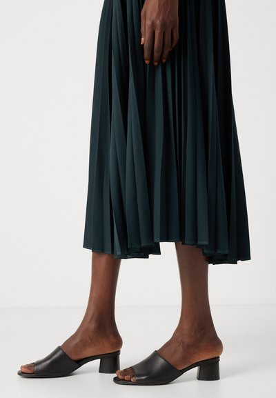Mules à talons en cuir noir avec un bout ouvert, associés à une jupe plissée vert foncé qui tombe au milieu du mollet et possède une texture lisse.