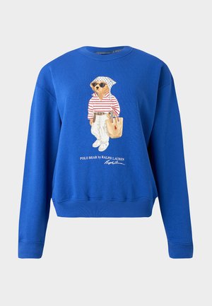 Γυναίκα που φοράει μπλε φούτερ με γραφικό αρκούδας, μαύρα αθλητικά παντελόνια και λευκά αθλητικά παπούτσια, στέκεται με τα χέρια πίσω από την πλάτη σε λευκό φόντο.