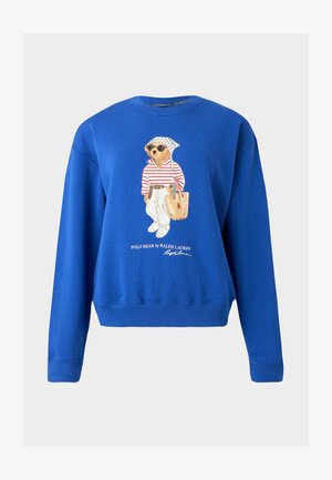 Blauwe trui met ronde hals met een beer die een zonnebril draagt, een gestreept topje, witte broek, een hoofddoek en een beige draagtas vasthoudt.