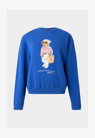 Μπλε φούτερ με στρογγυλή λαιμόκοψη με μια αρκούδα που φοράει γυαλιά ηλίου, ριγέ τοπ, λευκό παντελόνι, μαντίλι στο κεφάλι και κρατάει μια μπεζ τσάντα tote.