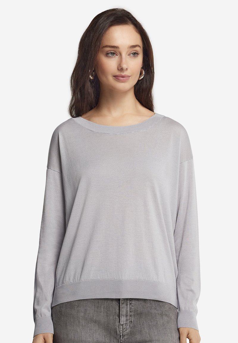 Jeune femme aux longs cheveux bruns portant un pull ample gris clair et un jean gris foncé, debout devant un fond uni.