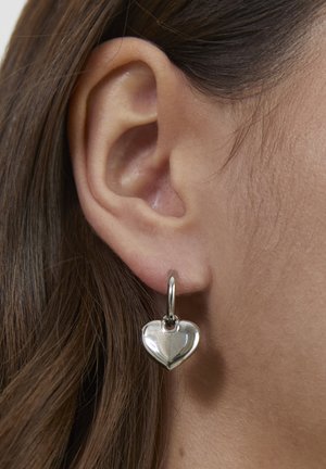 Silver heart-shaped earring with a polished finish, featuring a small hoop for attachment, shown near a human ear.