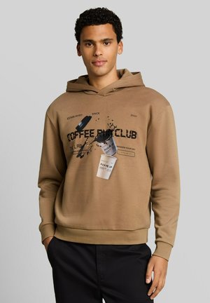 Junger Mann mit lockigem Haar trägt einen braunen Hoodie mit Abbildung einer Kaffeetasse und verschüttetem Kaffee sowie schwarze Hose, steht mit einer Hand in der Tasche.