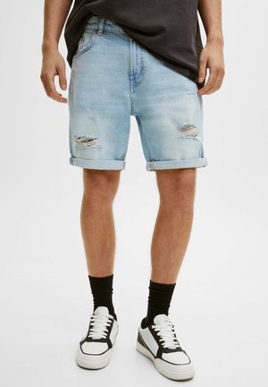Hombre vistiendo pantalones cortos de mezclilla desgastados y en azul claro con los dobladillos enrollados, calcetines negros y zapatillas blancas con detalles en negro, de pie contra un fondo liso.