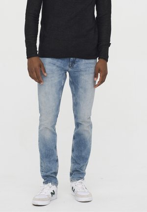 Mann trägt hellblaue Slim-Fit-Jeans, schwarzes Langarmshirt und weiße Sneakers, steht vor weißem Hintergrund.