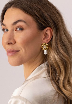 Frau mit welligem hellbraunem Haar, die ein cremefarbenes Satin-Oberteil und einen goldenen, blumenförmigen Ohrring mit Perlendetails trägt und über ihre Schulter schaut.