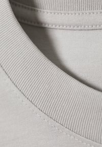 Materialul cămășii gri deschis are un guler ribbed și o textură netedă, cu cusături vizibile de-a lungul gulerului și corpului.