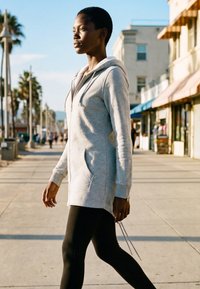 Junge Frau in grauem Hoodie und schwarzen Leggings, die entlang eines sonnigen Bürgersteigs mit Palmen und Geschäften sowie Gebäuden im Hintergrund geht.