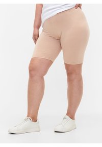 Person wearing beige fitted knee-length shorts and white sneakers, standing against a plain white background.