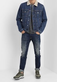 Chaqueta de mezclilla con un cuello de pelaje sintético crema, camiseta gris, jeans desgastados azul y zapatillas verdes olivo con suelas blancas.