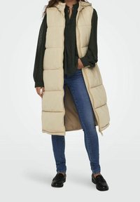 Beige gepolsterte Weste mit Kapuze, ausgestattet mit einem Reißverschluss und einem langen Design. Kombiniert mit einer dunkelgrünen Bluse, blauen Jeans und schwarzen Slippern.