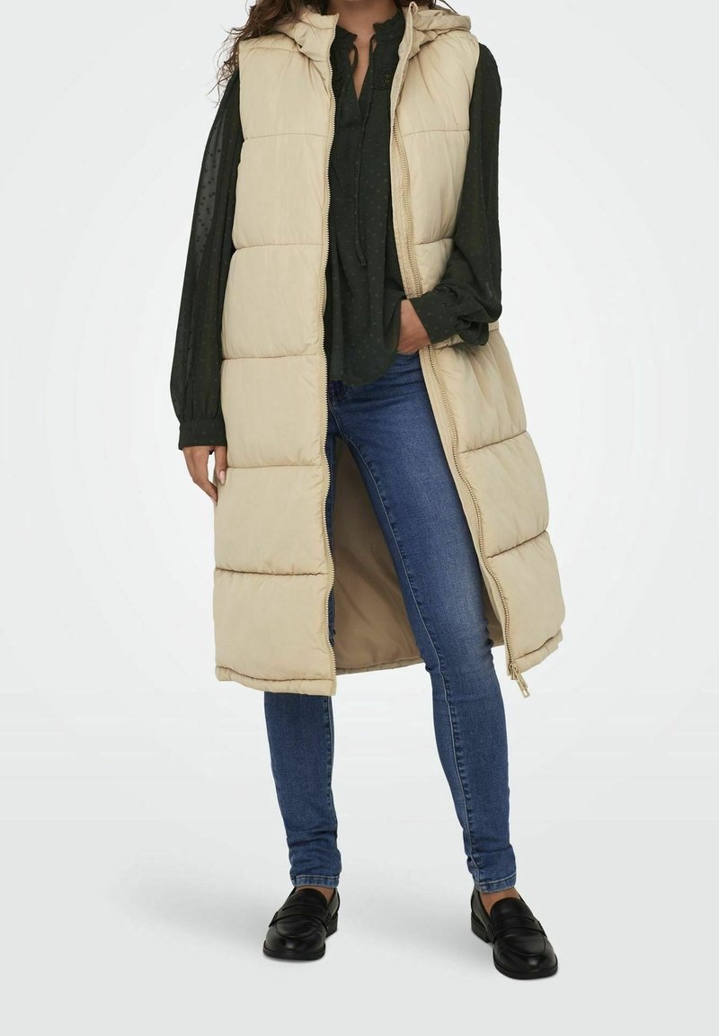Beige gepolsterte Weste mit Kapuze, ausgestattet mit einem Reißverschluss und einem langen Design. Kombiniert mit einer dunkelgrünen Bluse, blauen Jeans und schwarzen Slippern.