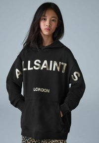 Sudadera negra con texto metálico dorado "ALLSAINTS" y "LONDRES". Tiene un bolsillo frontal, capucha y puños de canalé. Se llevan pantalones de leopardo por debajo.