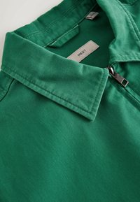 Veste à col vert avec une fermeture éclair argentée, montrant l'étiquette intérieure avec le nom de la marque "NEXT".