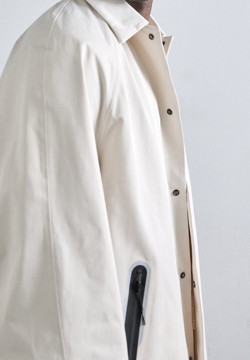 Osoba ubrana w beżowy płaszcz przeciwdeszczowy z czarnym zamkiem na kieszeni, stojąca na tle jednolitego jasnoszarego tła.