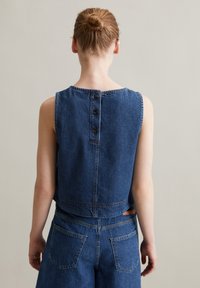 Jeansowy top bez rękawów z okrągłym dekoltem, widocznymi szwami i trzema guzikami na plecach; w komplecie z dżinsowymi spodniami o wysokim stanie.