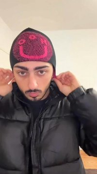 Černá čepice s růžovým usmívajícím se obličejem, vyrobená z elastické látky, nošená s černou nadýchanou bundou. Pozadí je neutrální.