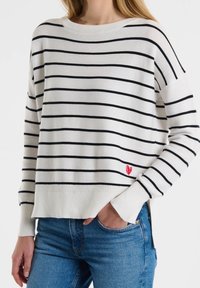 Weißer Pullover mit schwarzen Streifen, geripptem Rundhalsausschnitt und Bündchen, mit einem kleinen roten Herzakzent am Saum. Kombiniert mit blauen Jeans.