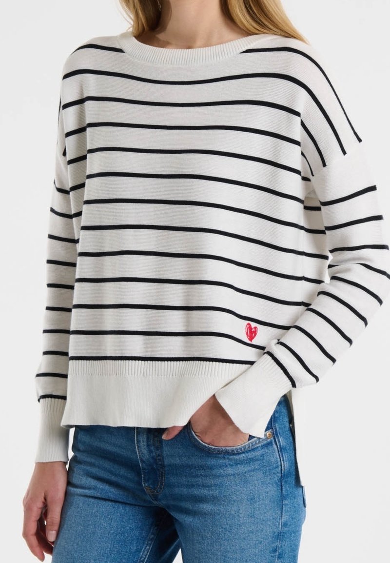 Weißer Pullover mit schwarzen Streifen, geripptem Rundhalsausschnitt und Bündchen, mit einem kleinen roten Herzakzent am Saum. Kombiniert mit blauen Jeans.
