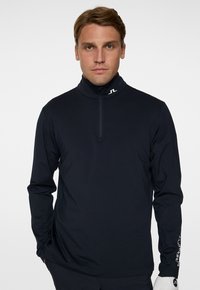 Pullover nero a maniche lunghe con colletto alto, zip sul davanti e logo sul collo. Tessuto liscio, design aderente con aperture per il pollice sui polsini.