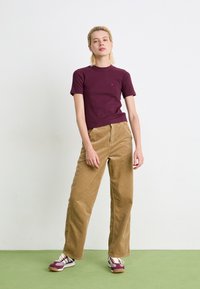 Camicia a maniche corte Marlowe in bordeaux, abbinata a pantaloni larghi in corduroy color beige e scarpe da ginnastica color marrone con accenti bianchi.